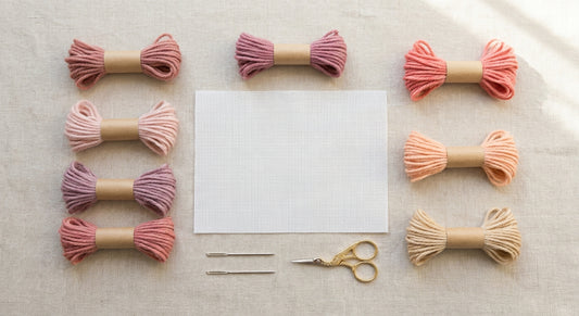 Needlepoint canvas surrounded by seven pink wool shades, needles, and scissors