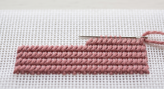 Continental stitch rows in dusty rose wool on white 13-mesh needlepoint canvas, with tapestry needle mid-row
